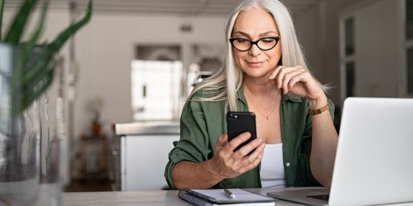 Kvinne sitter foran pc-en og sjekker mobilen